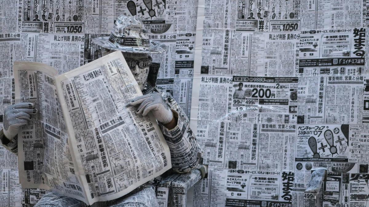 Image depicting a man reading a newspaper dressed in a full suit made of newspaper print, standing in front of a wall completely plastered with newspaper pages.