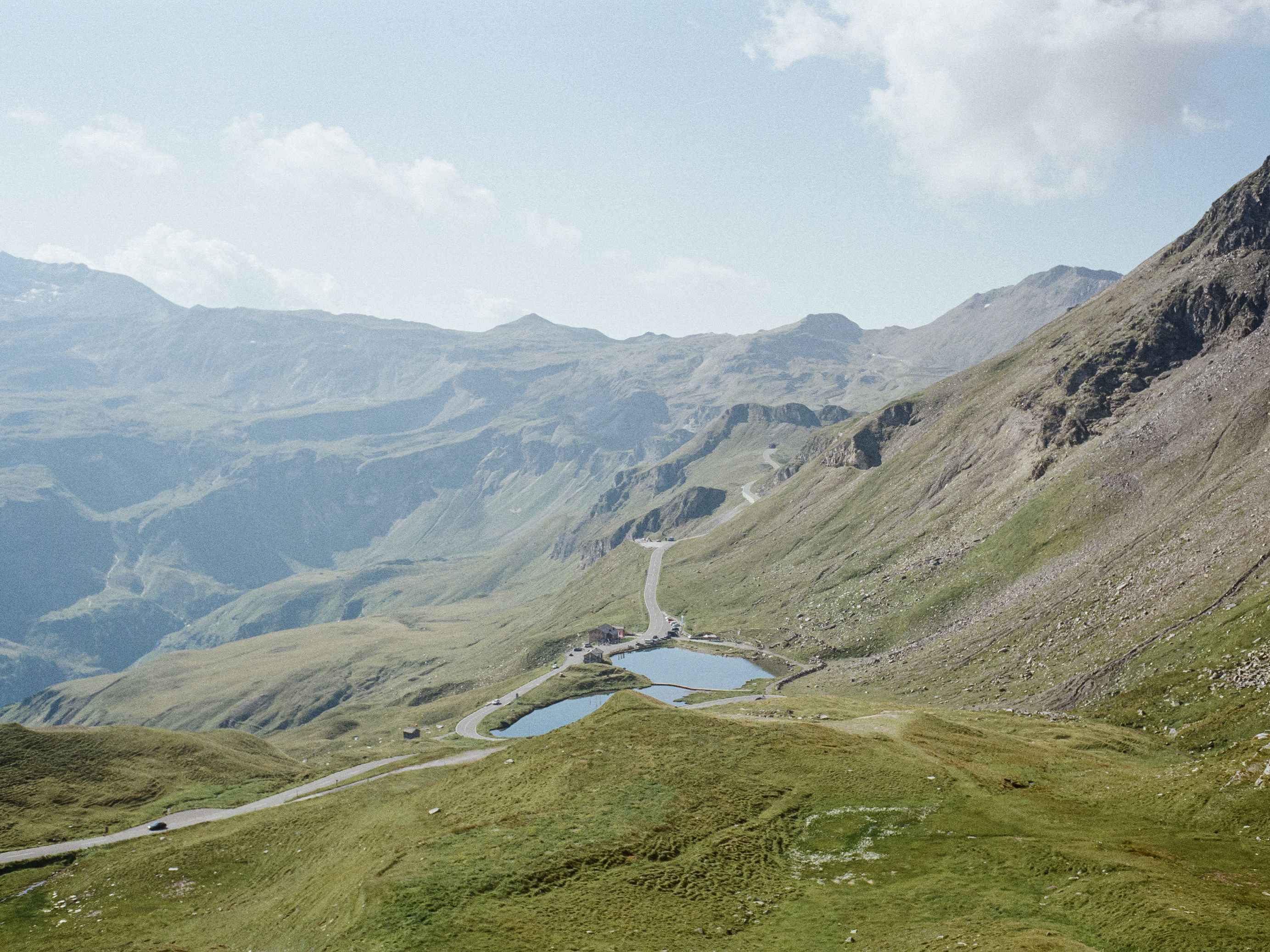 a sunny day on the Grossglockner, between the ridges of rock and grass is a road winding past a remote hut