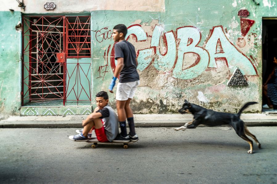 Joe Willems Photography Cuba Niños en monopatín y perro saltando frente a una pared turquesa