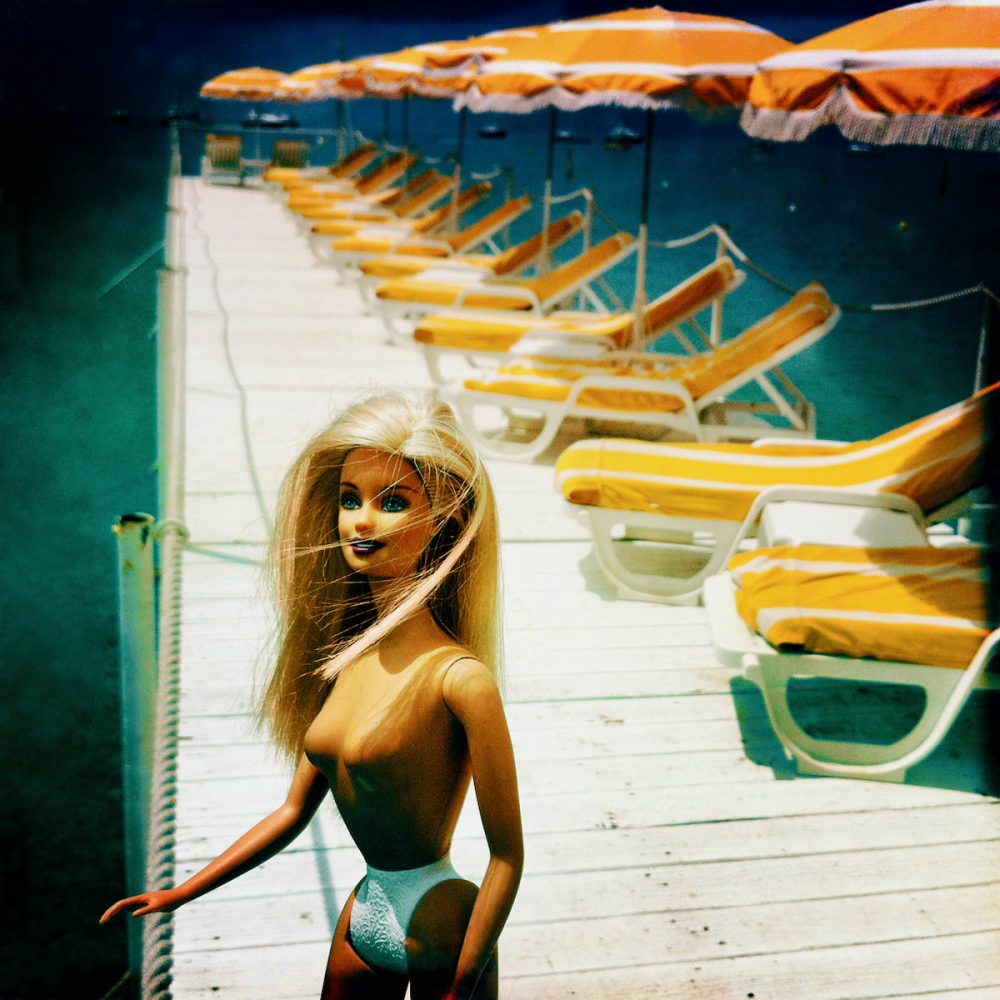 Delia Dickmann Fotografie nackte blonde Barbie an einem Steg mit gelben Sonnenliegen