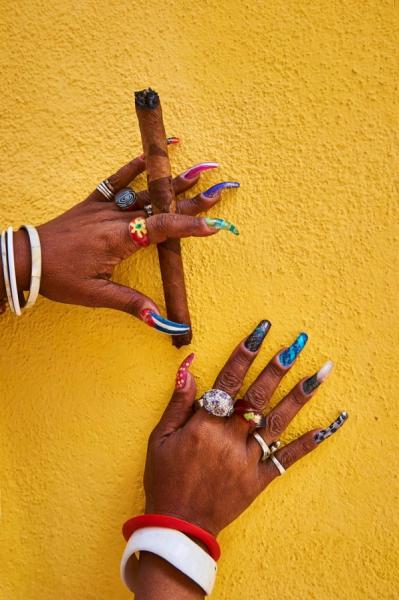 Joe Willems Fotografie Hände einer afrikanischen Frau mit langen Acryl Nägeln die eine Zigarre hält vor gelber Wand