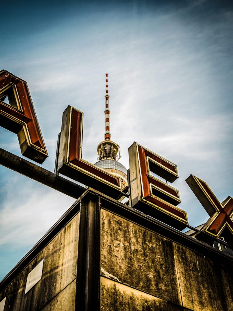 Georgia Ortner Fotografie perspektivisch Alexanderplatz Schild mit blauem Himmel und Berliner Fernsehturm im Hintergrund