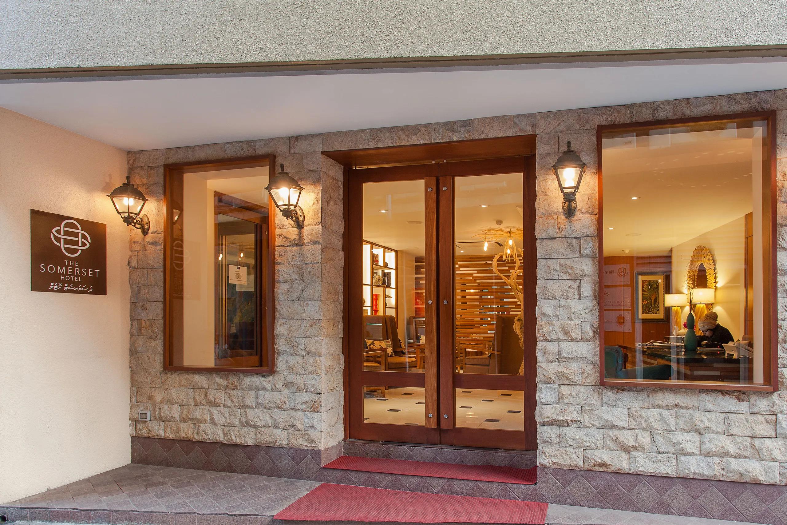 Stone-clad street entrance of The Somerset Hotel in Malé at dusk, with lantern lights, wooden double doors and the hotel sign