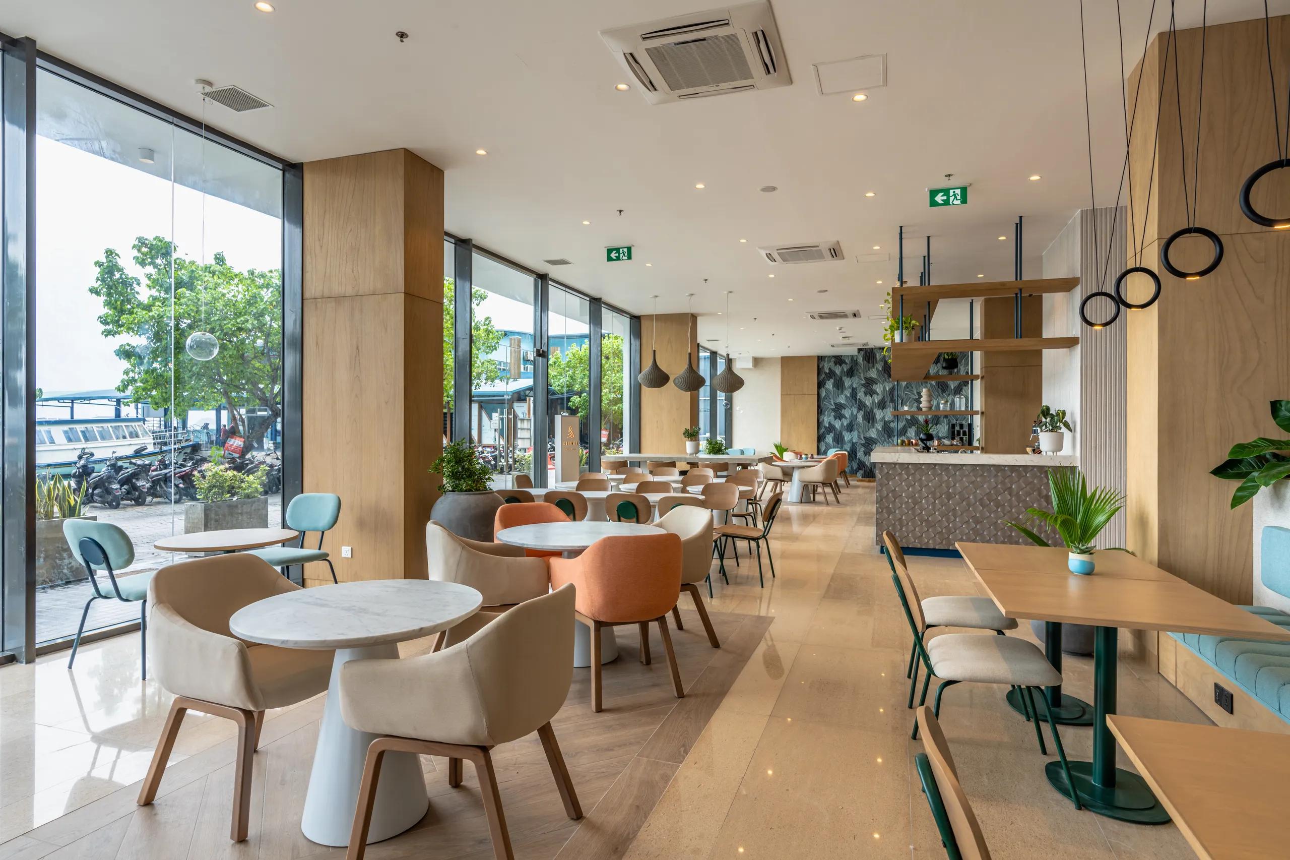Ground-floor interior of Alimas Cafe & Lounge with pastel armchairs, round marble tables and floor-to-ceiling windows onto the Malé waterfront