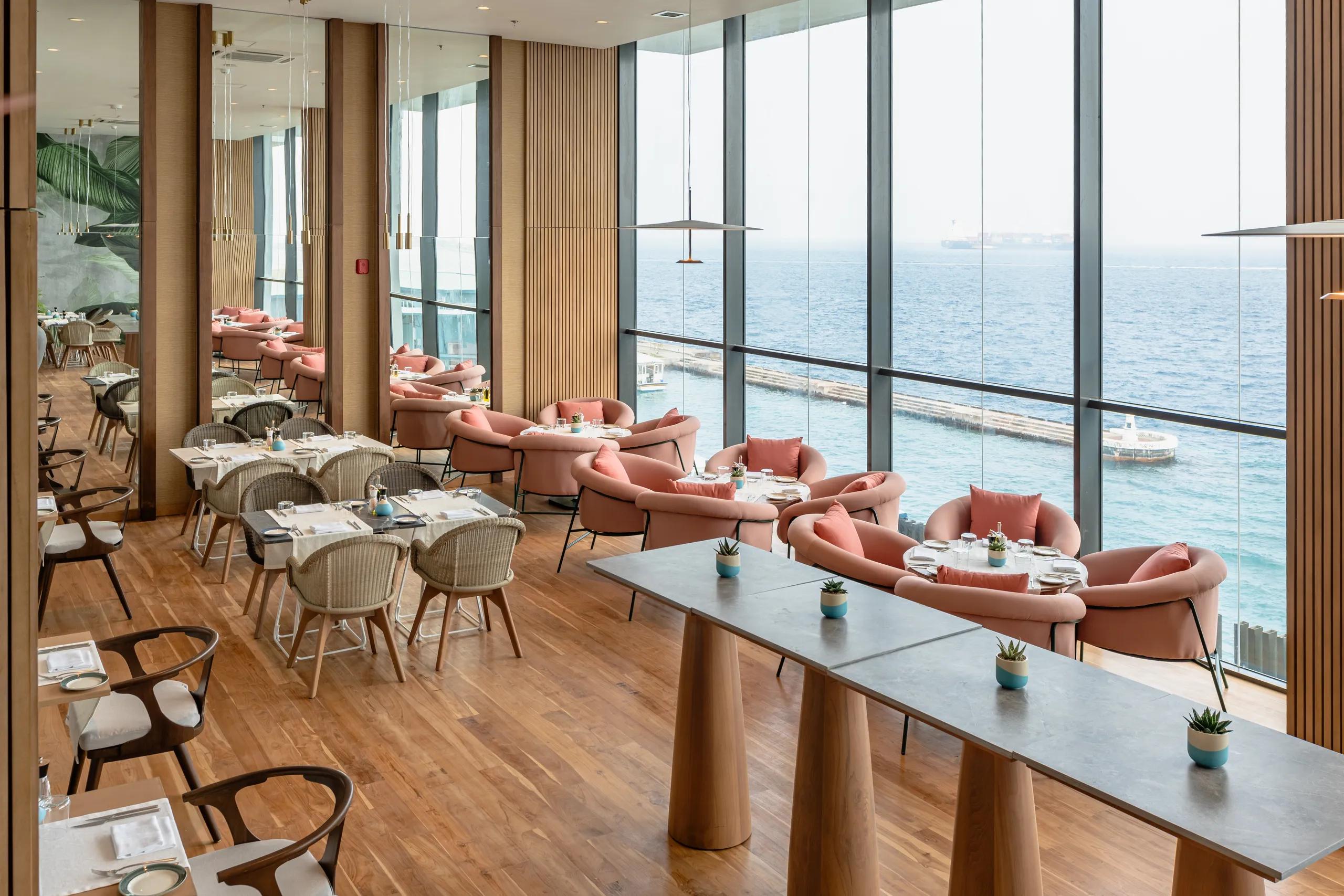 Dining room of Oivaru Restaurant with pink armchairs, laid tables and floor-to-ceiling windows overlooking the sea off Malé