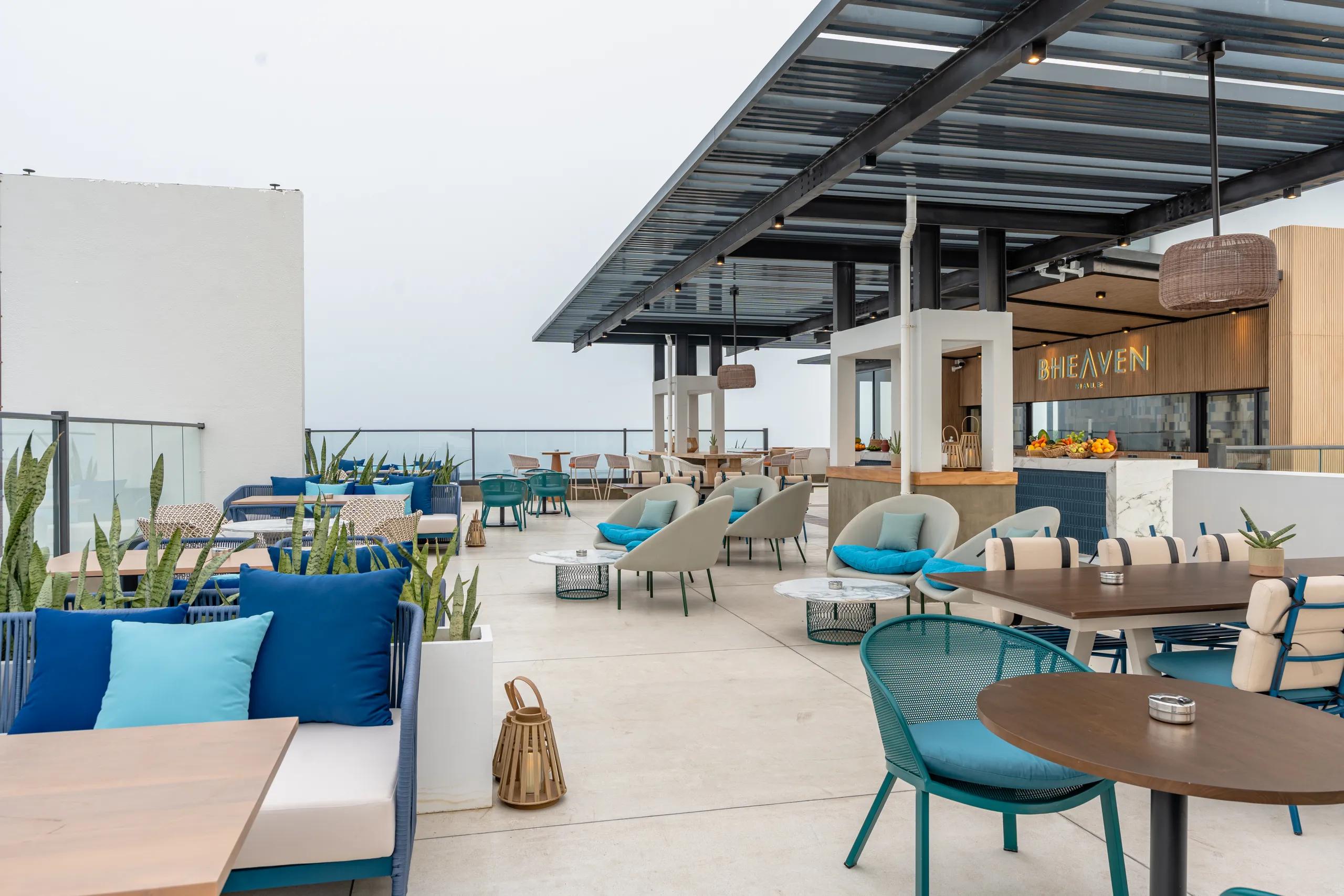 Open-air rooftop terrace of B.Heaven with blue lounge seating under a pergola and the lit B.Heaven sign above the bar counter