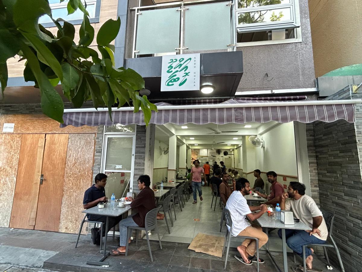 Addu Hotaa's open front under a striped awning, with its green Thaana sign above and diners at pavement tables