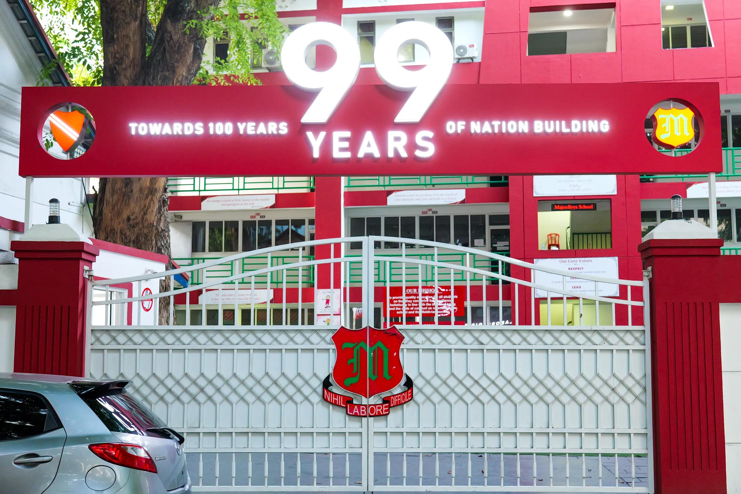 The red and white gate of Majeedhiyya School in Malé, with its 99 Years of Nation Building banner and the school crest