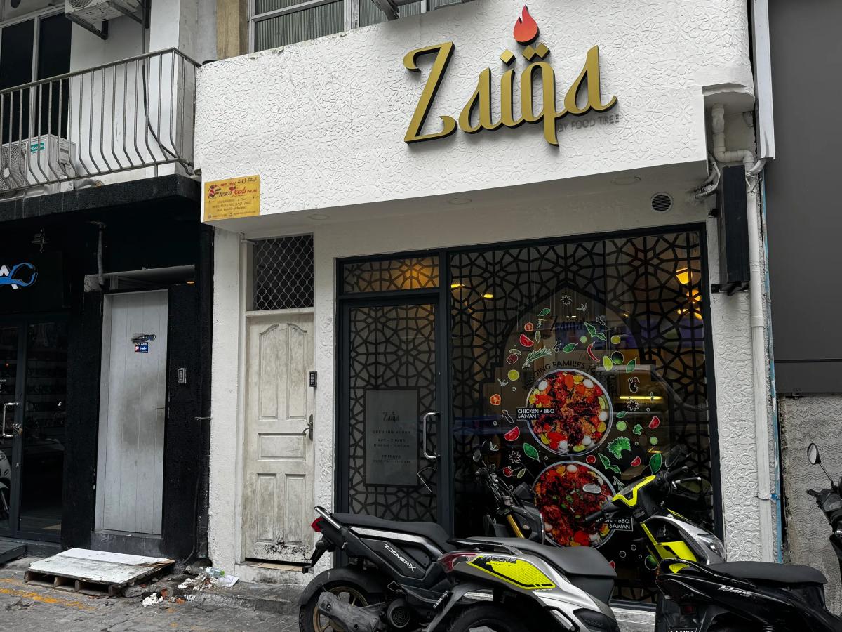 Gold Zaiqa sign above a glass storefront with patterned screen and food graphics, motorbikes parked in front