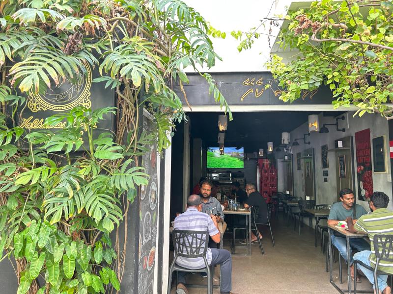 Open-fronted Valhomas Cafe with men seated at tables, a wall-mounted TV and dense greenery at the entrance