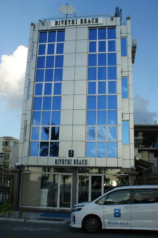 Street front of Rivethi Beach Hotel in Hulhumalé, a white building with blue glass windows and the hotel's branded van outside