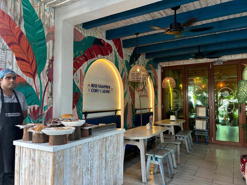 Tropical-themed interior at The Red Snapper & Coffee Beans with a pastry display, wooden tables and a leaf mural