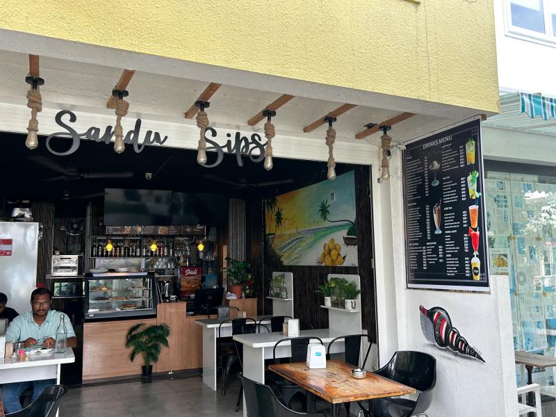 Sandy Sips cafe with cursive sign, hanging rope lights, a beach mural, drinks menu board and outdoor tables
