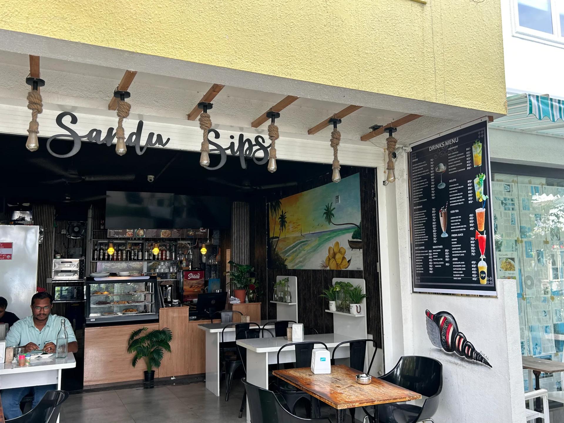 Sandy Sips cafe with cursive sign, hanging rope lights, a beach mural, drinks menu board and outdoor tables