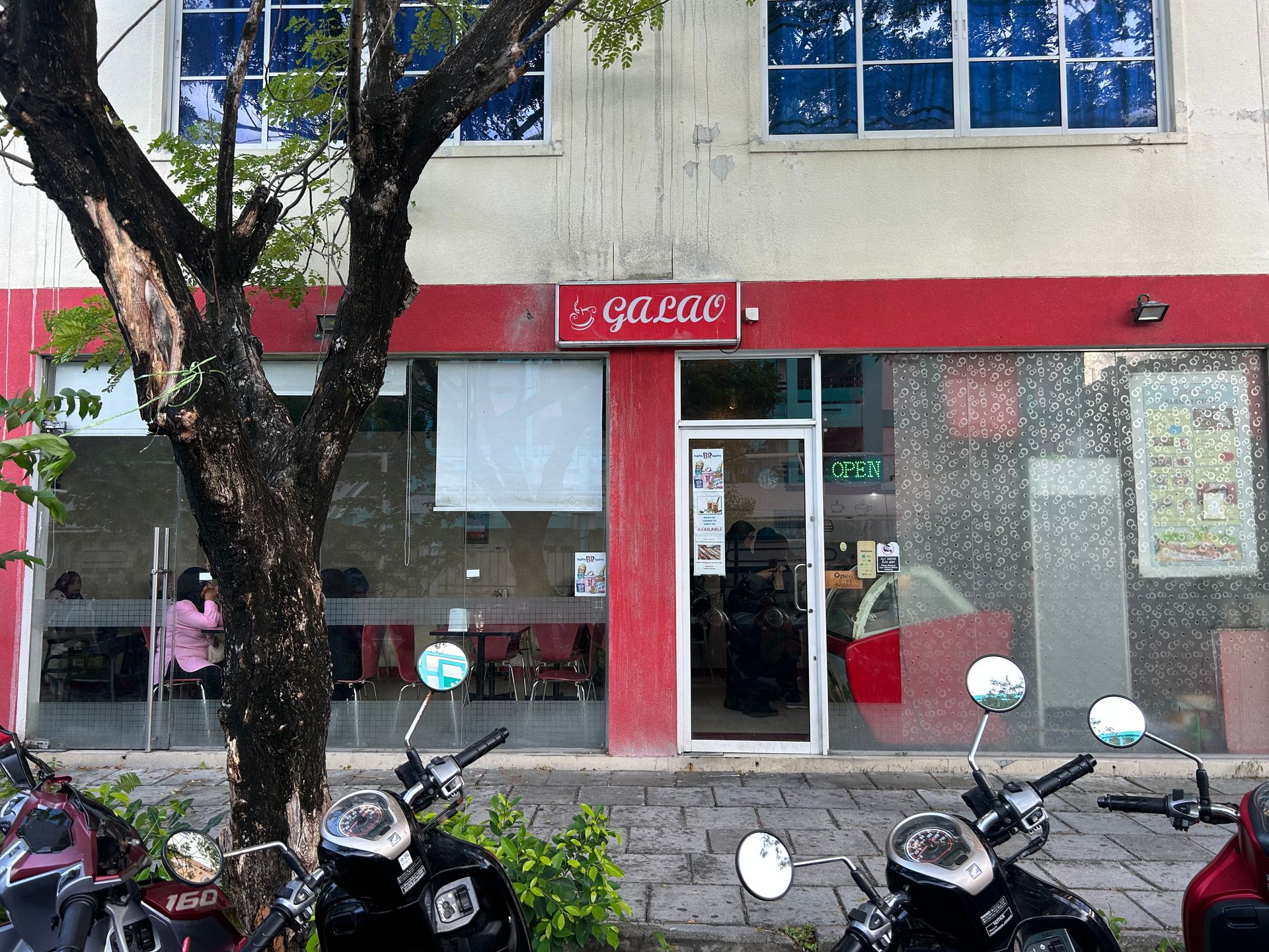 Red Galao cafe storefront with an Open sign, seen past a tree and parked scooters