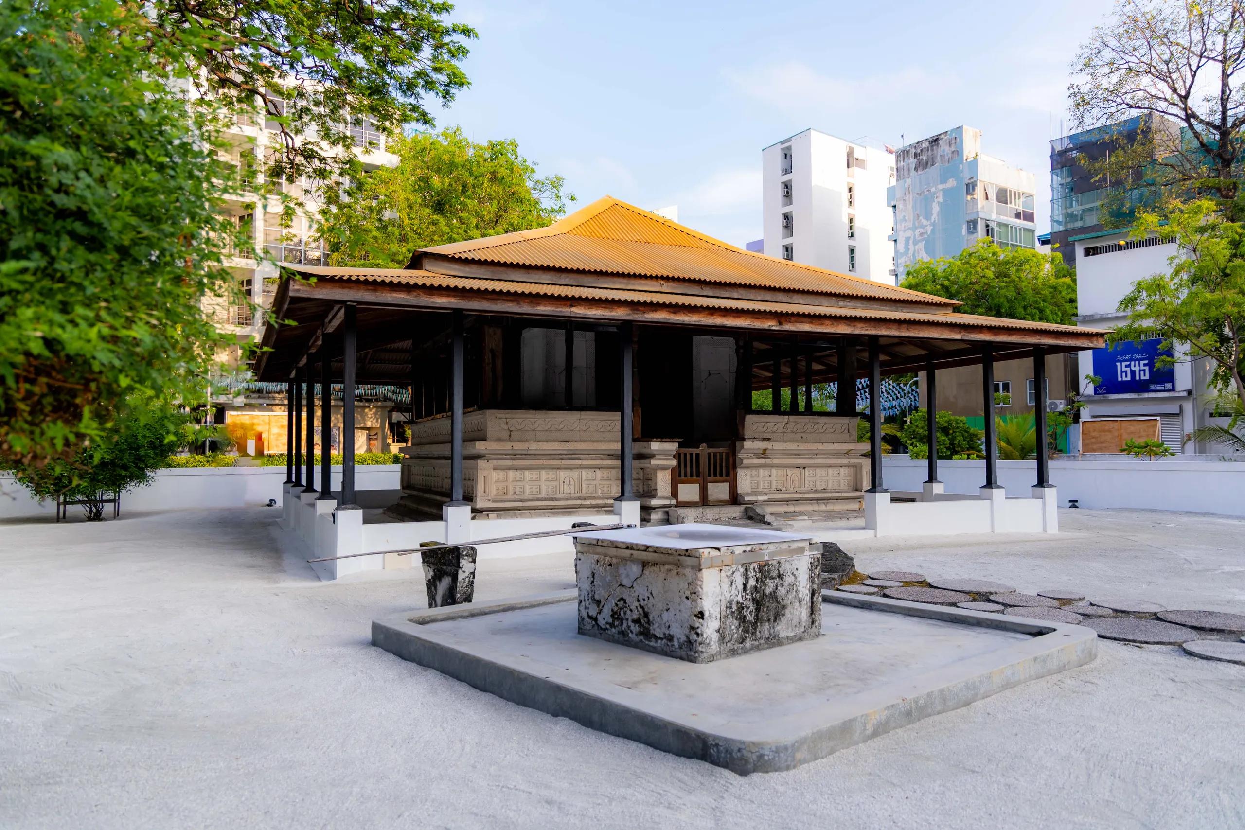 Open pavilion with a tiled roof sheltering carved coral-stone tombs at Kalhuvakaru Miskiy, city buildings behind