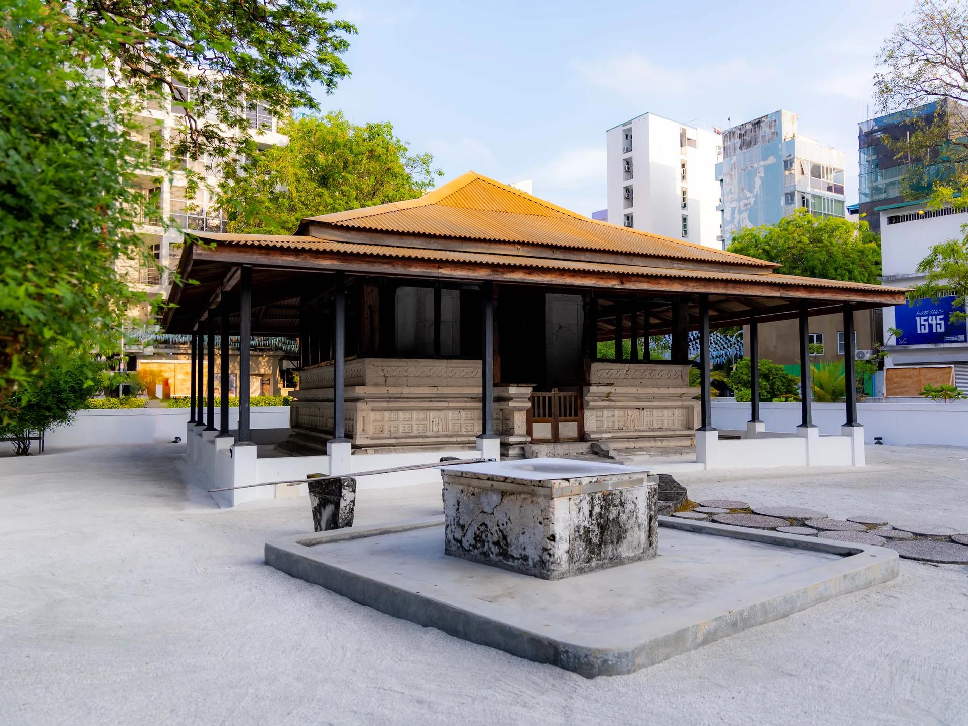 Open pavilion with a tiled roof sheltering carved coral-stone tombs at Kalhuvakaru Miskiy, city buildings behind