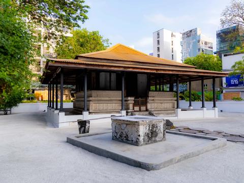 Open pavilion with a tiled roof sheltering carved coral-stone tombs at Kalhuvakaru Miskiy, city buildings behind
