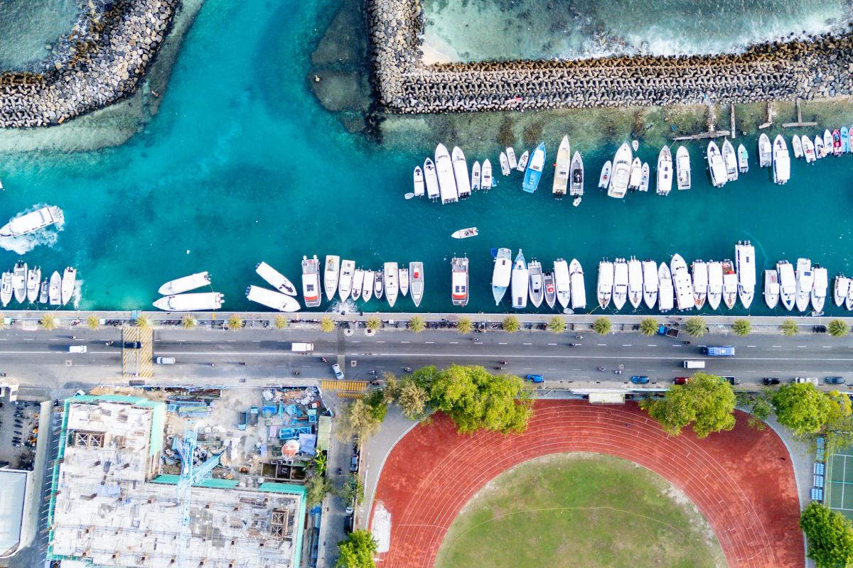 Top-down drone view of Malé's harbour front, with speedboats moored along the teal water, the palm-lined waterfront road and the running track beside it