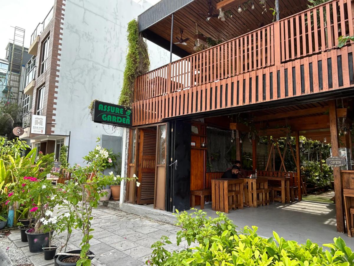 Assure Garden's green sign beside a timber deck, with wooden tables in the plant-filled open dining area