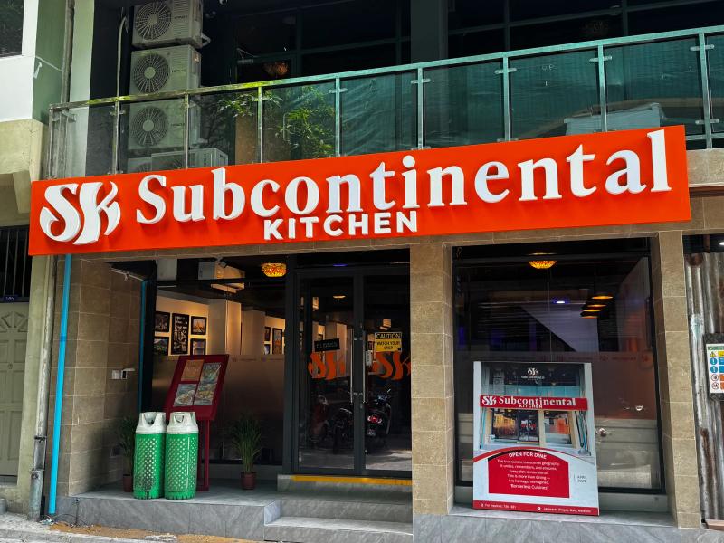 Orange Subcontinental Kitchen sign above a glass-fronted restaurant entrance with gas cylinders by the door