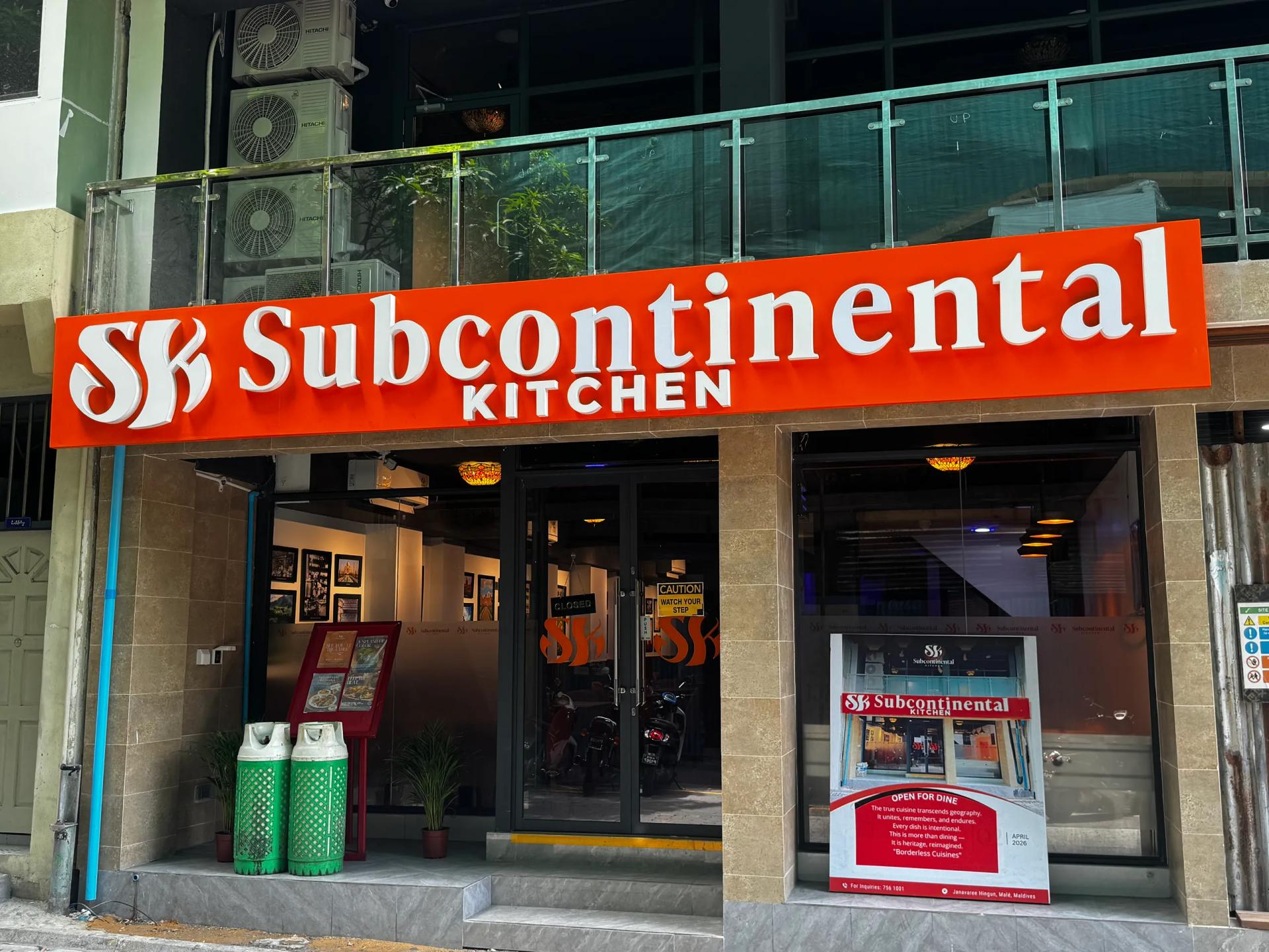 Orange Subcontinental Kitchen sign above a glass-fronted restaurant entrance with gas cylinders by the door