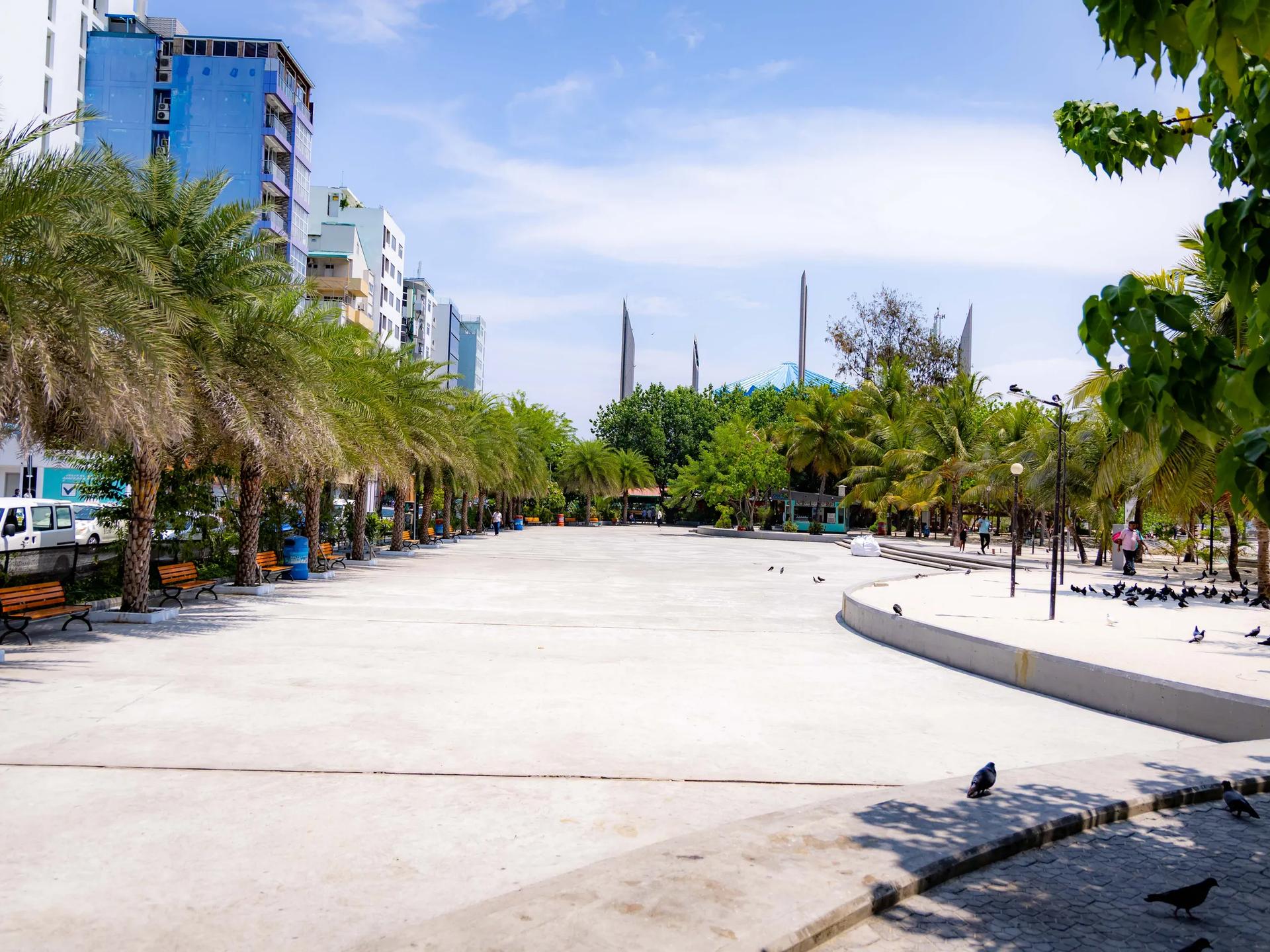 Wide paved plaza lined with palms and benches near Artificial Beach, mosque spires and apartment blocks behind