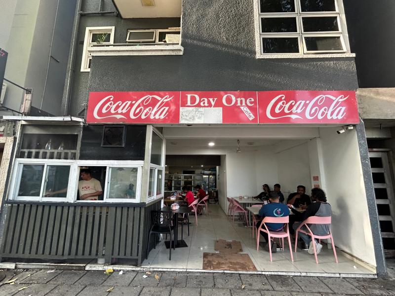 Day One Cafe's entrance under a red Coca-Cola sign band, with pink chairs in the open dining area