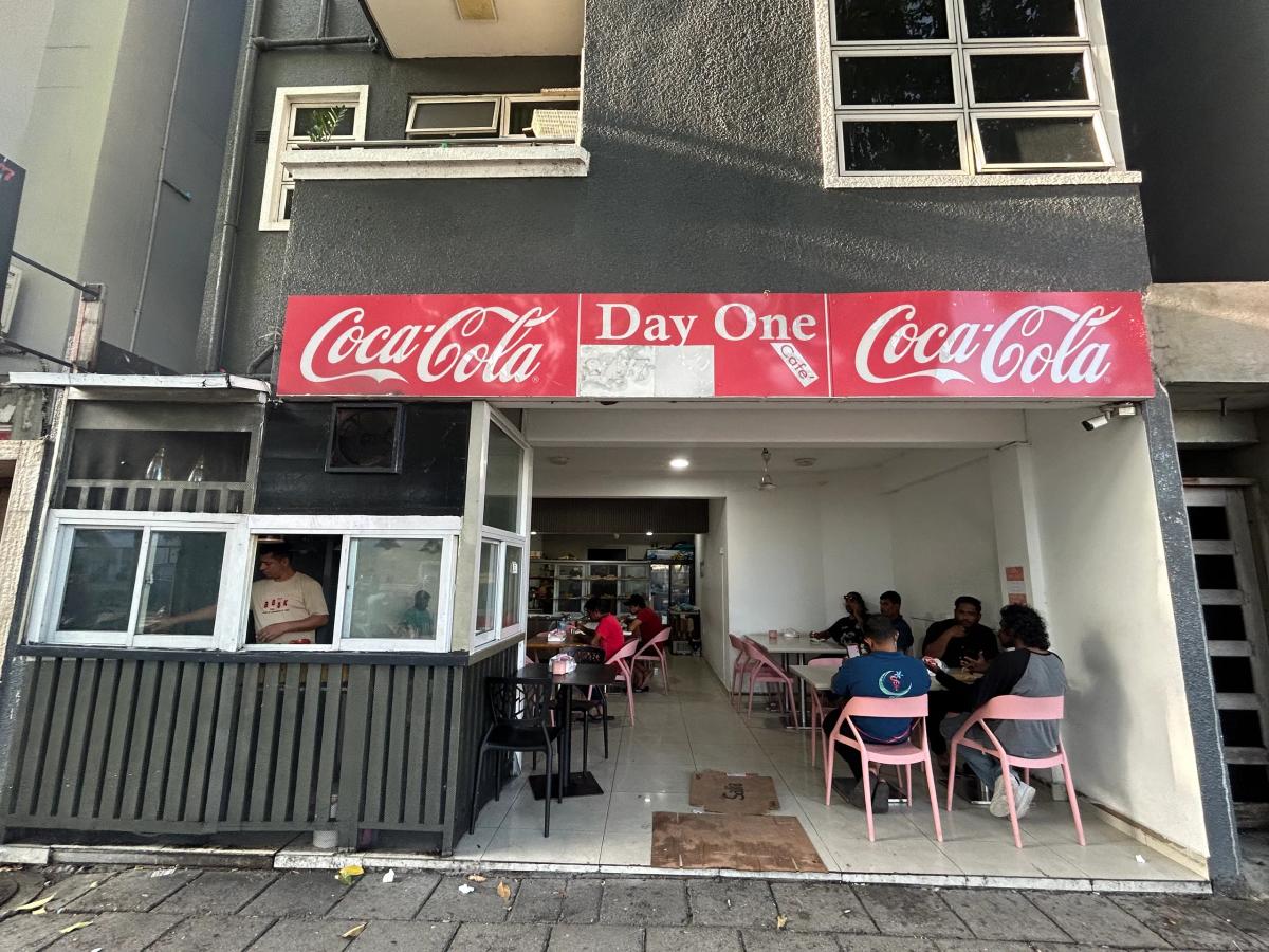 Day One Cafe's entrance under a red Coca-Cola sign band, with pink chairs in the open dining area