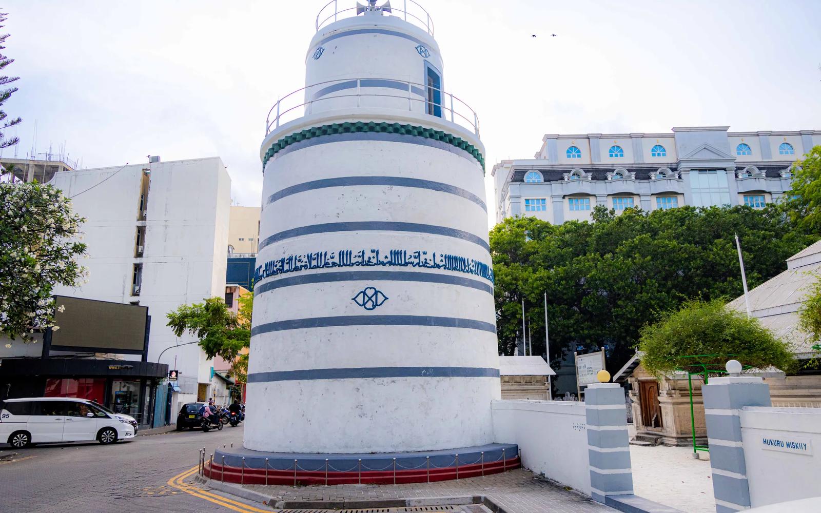 White and blue cylindrical minaret of Hukuru Miskiy, with a sign reading Hukuru Miskiy on the wall beside it