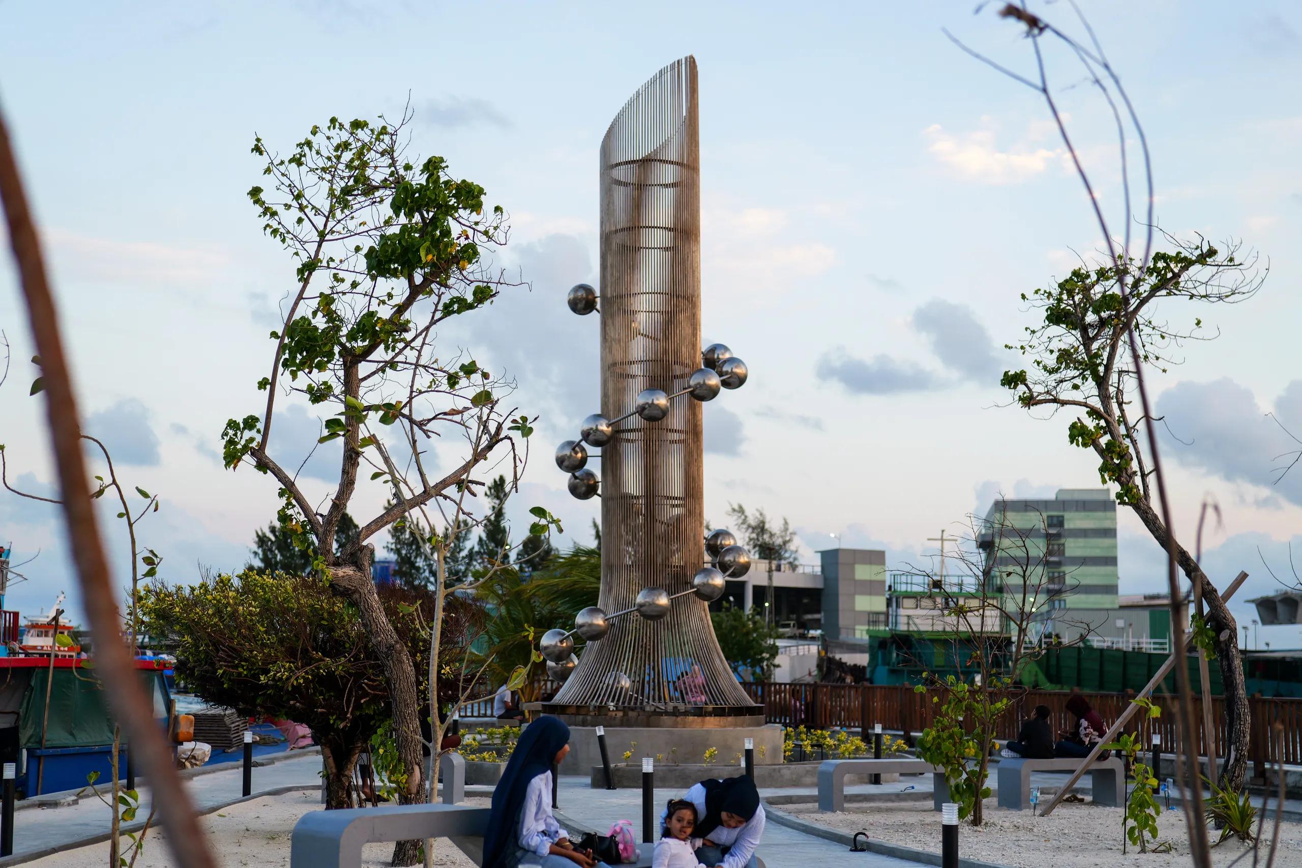 The steel Tsunami Monument, a spiral of metal spheres around a column of vertical rods, in a landscaped seafront plaza at dusk