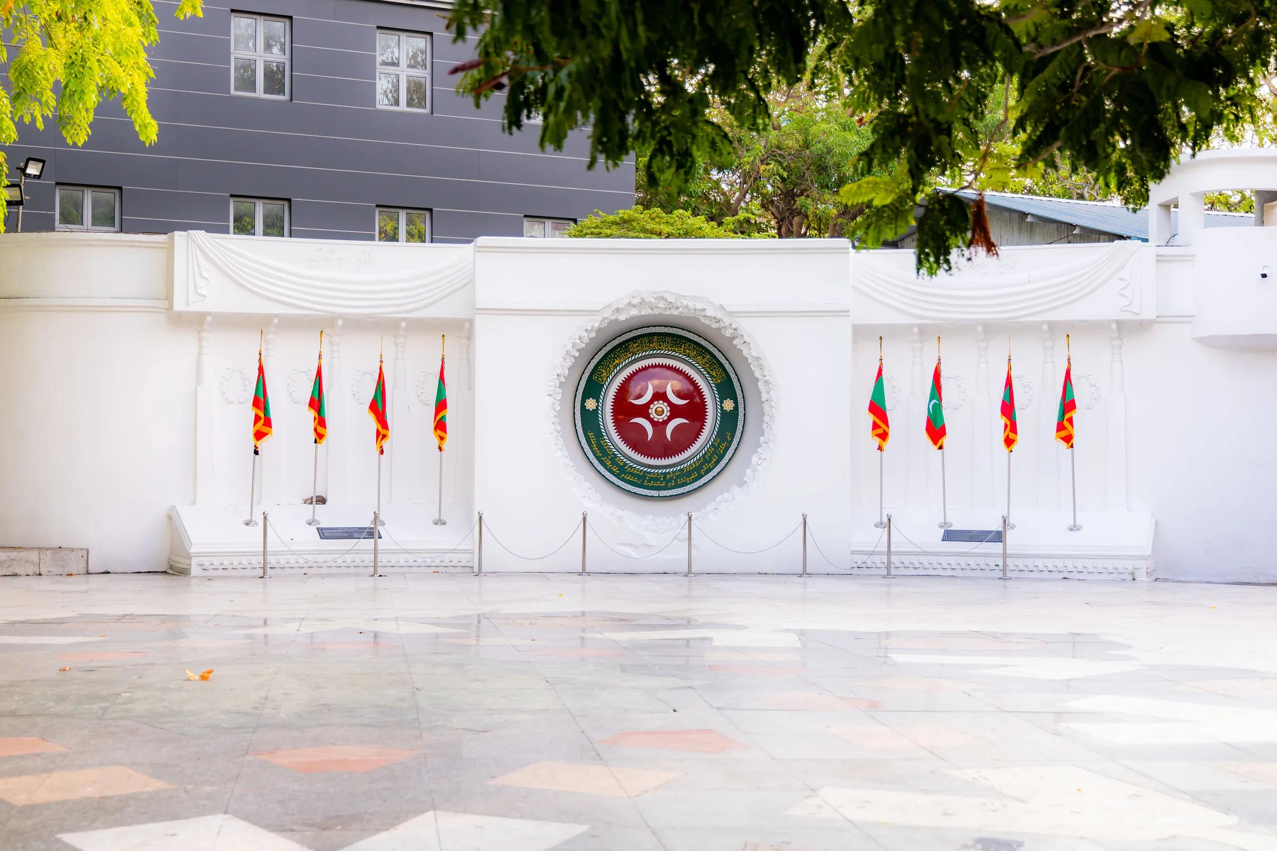 White Victory Monument wall with the Maldives national emblem and rows of flags on a tiled plaza