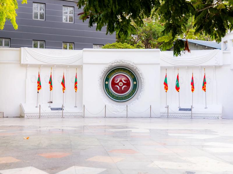 White Victory Monument wall with the Maldives national emblem and rows of flags on a tiled plaza