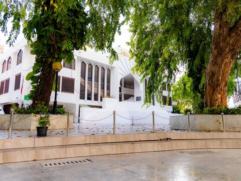 White facade of the Islamic Centre with tall arched windows, shaded by large trees over a paved forecourt