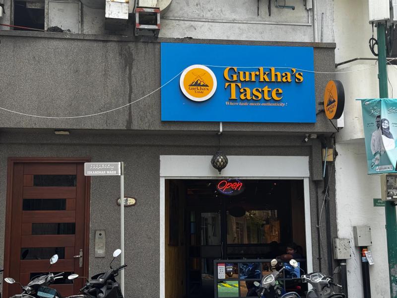 Blue Gurkha's Taste sign on a grey building with a neon Open sign over the doorway on Iskandhar Magu