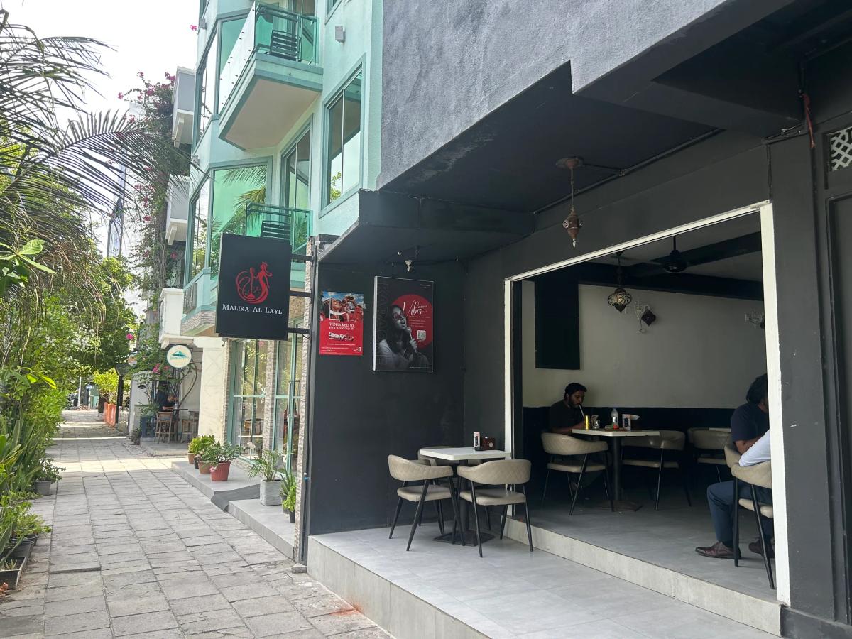 Malika Al Layl hanging sign and an open dark-walled dining area with cushioned chairs beside a Malé sidewalk