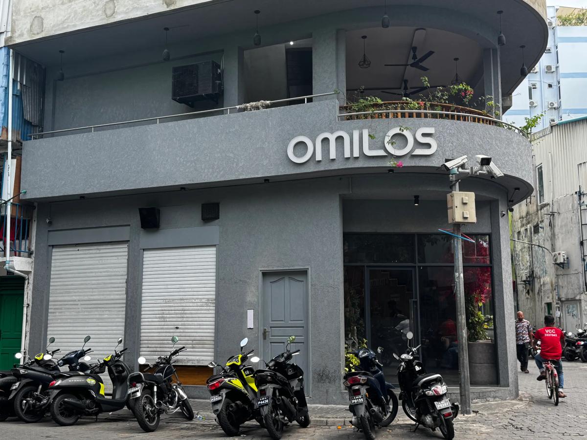 Grey corner building with Omilos signage and an upper rooftop terrace, motorbikes parked along the street