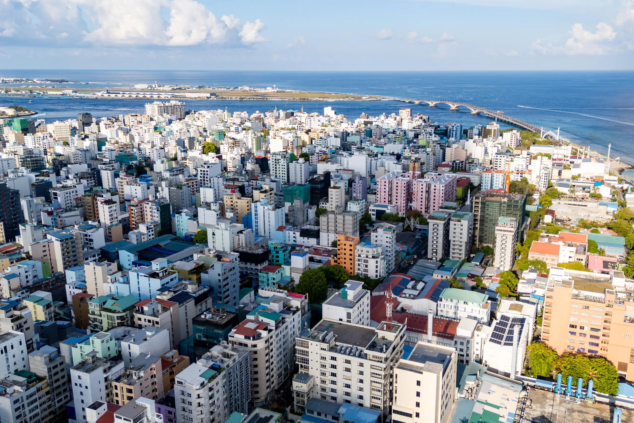 Aerial view over the dense rooftops of Malé, with the Sinamalé Bridge curving across the channel toward Hulhulé on the horizon