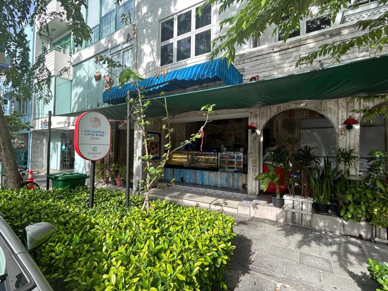 Street-side takeaway counter of The Red Snapper & Coffee Beans Express in Hulhumalé, with a weathered-wood facade, a round logo sign reading Takeaway, a glass pastry display case and potted plants behind a low hedge