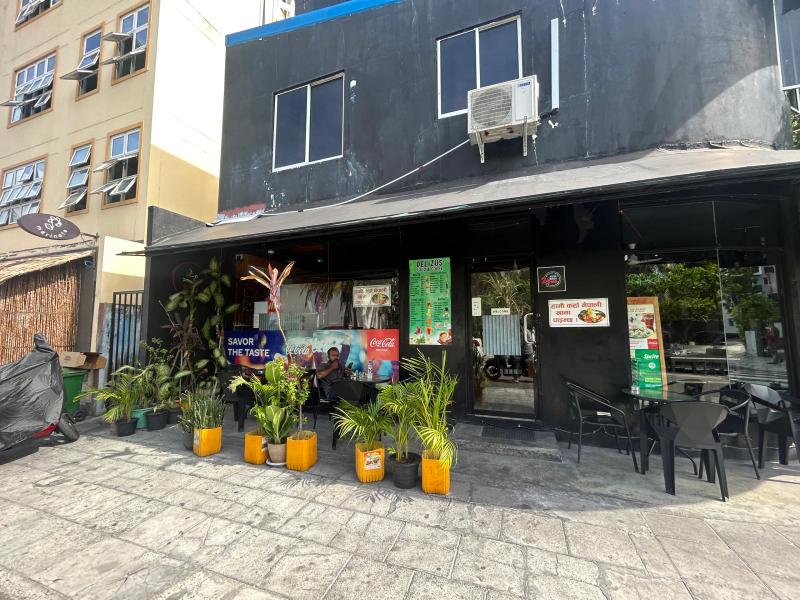 Delizus Food & Coffee's black corner storefront with menu posters, potted plants and pavement tables