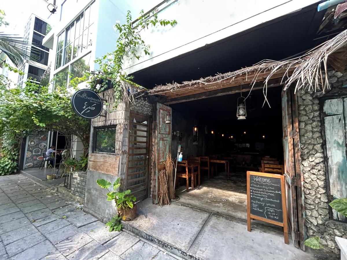 The Chef Mode Badhige rustic entrance with stone walls, thatched roof, wooden tables and a Welcome chalkboard