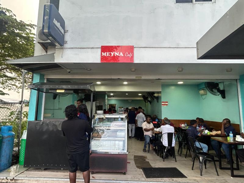 Open-fronted Meyna Cafe with a red sign, a glass display case and diners at tables in the turquoise interior
