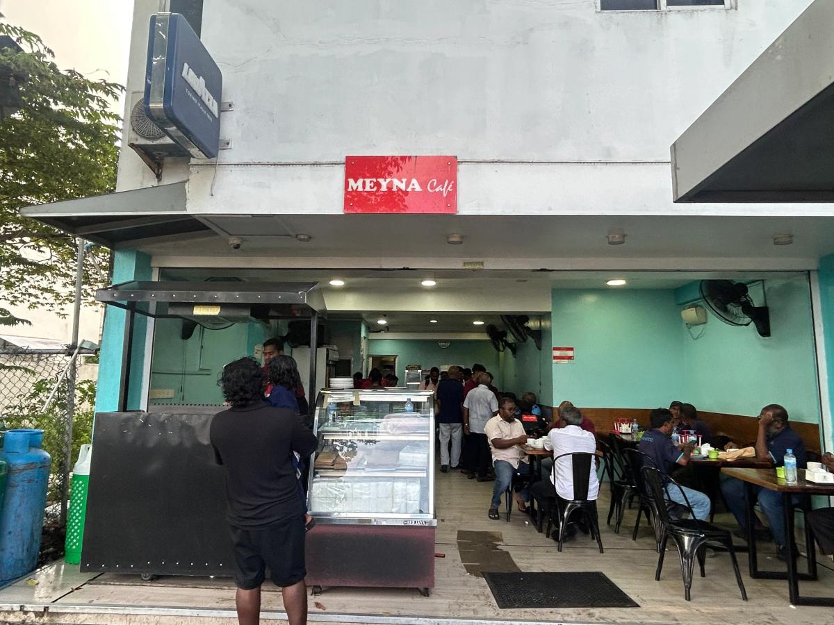 Open-fronted Meyna Cafe with a red sign, a glass display case and diners at tables in the turquoise interior