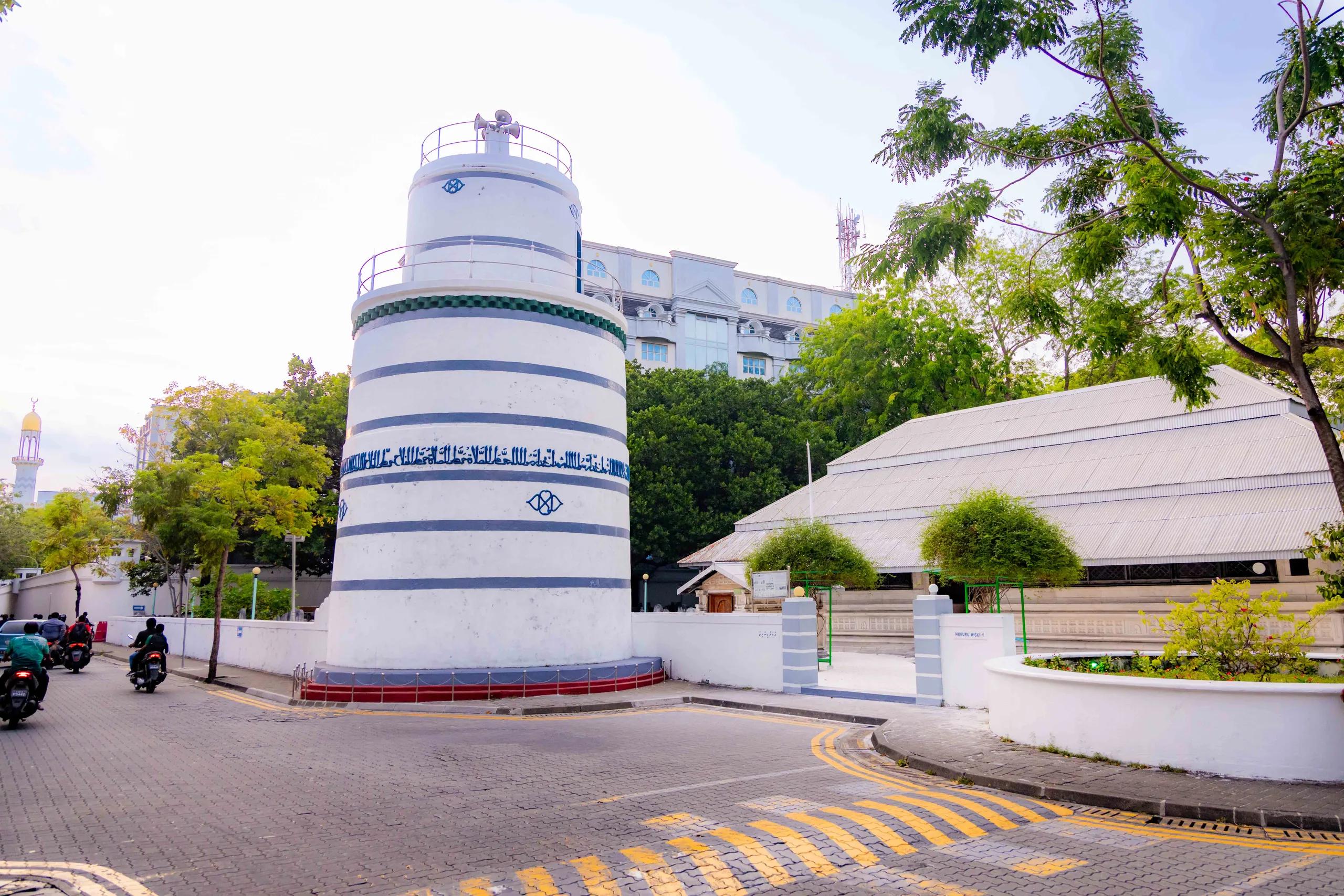 The white and blue Munnaaru minaret in Malé with motorbikes passing on the street, the roof of Hukuru Miskiy beside it and the gold dome of the Islamic Centre in the distance