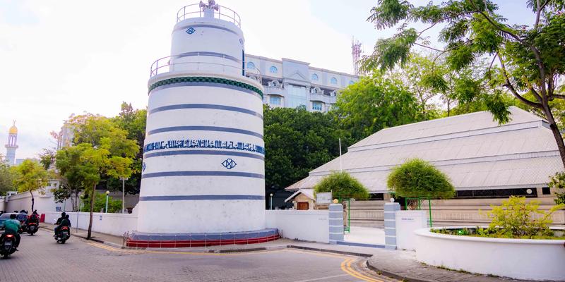 The white and blue Munnaaru minaret in Malé with motorbikes passing on the street, the roof of Hukuru Miskiy beside it and the gold dome of the Islamic Centre in the distance