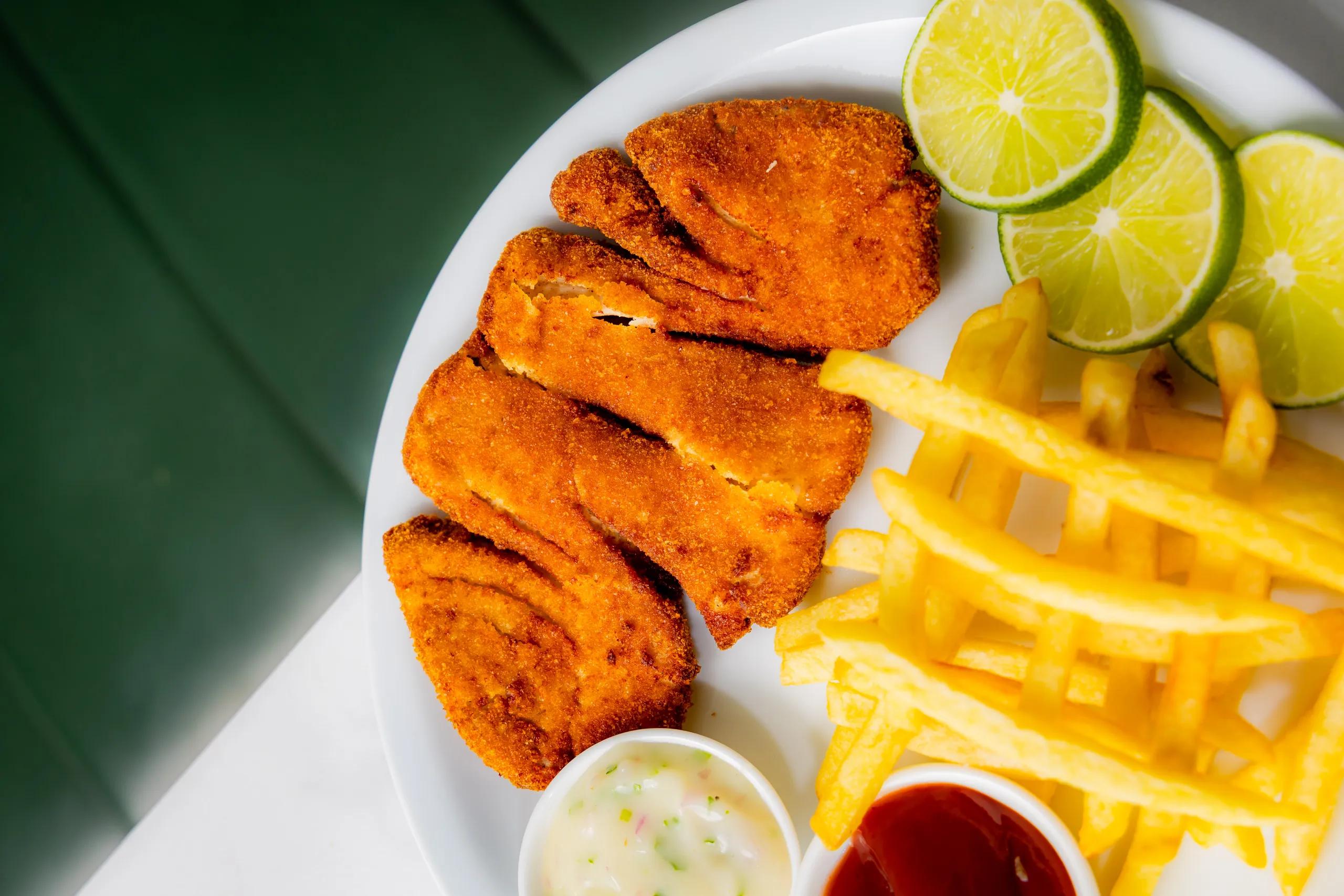 Breaded fried fish fillets with french fries, lime slices and dipping sauces on a white plate