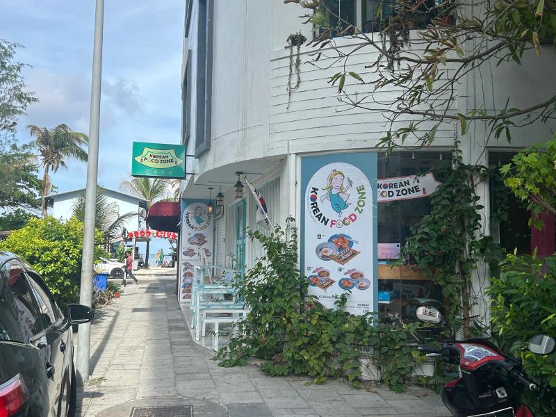 Korean Food Zone sign and illustrated menu banners on a white street-corner building, with a sidewalk and palm trees