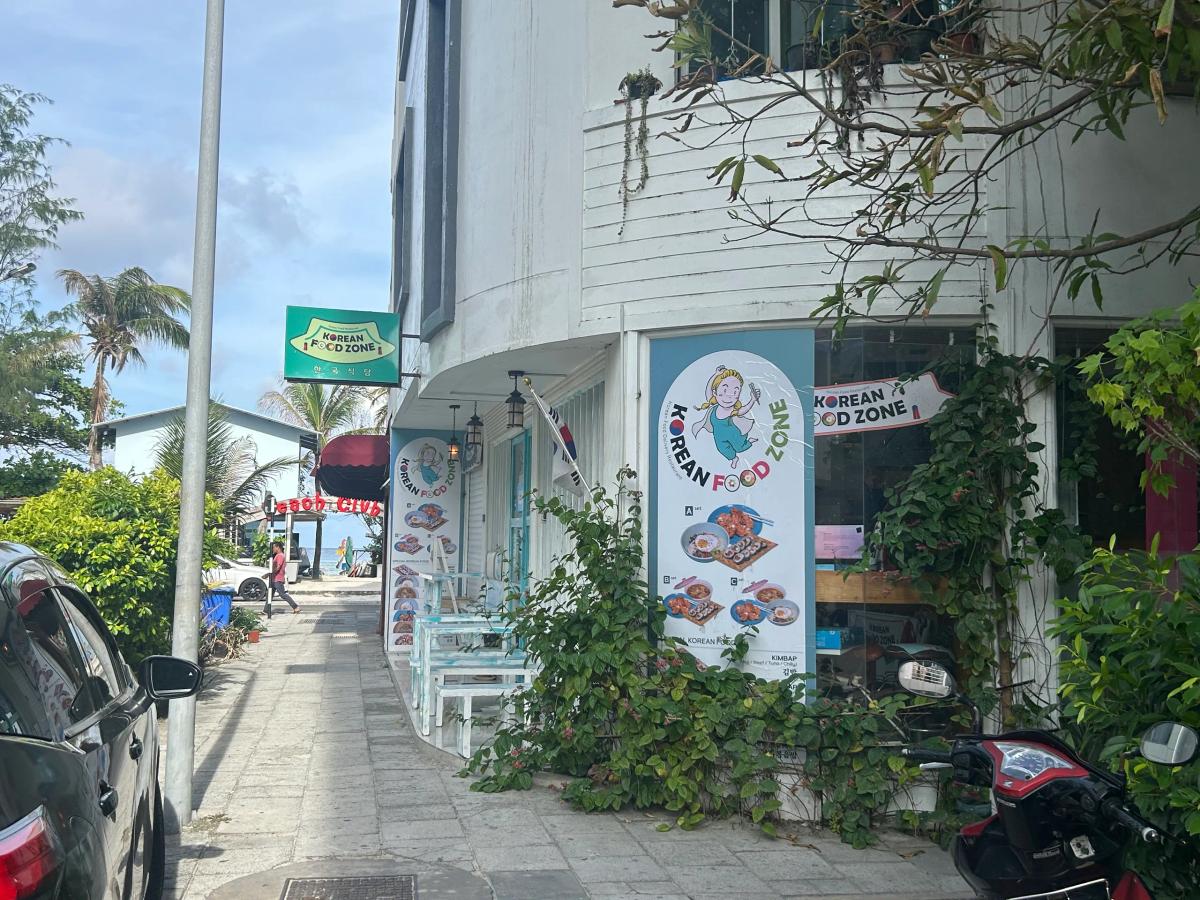 Korean Food Zone sign and illustrated menu banners on a white street-corner building, with a sidewalk and palm trees