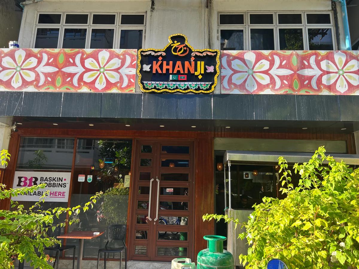 Glowing Khanji sign with floral mural above the glass storefront of Khanji restaurant, with leafy plants out front