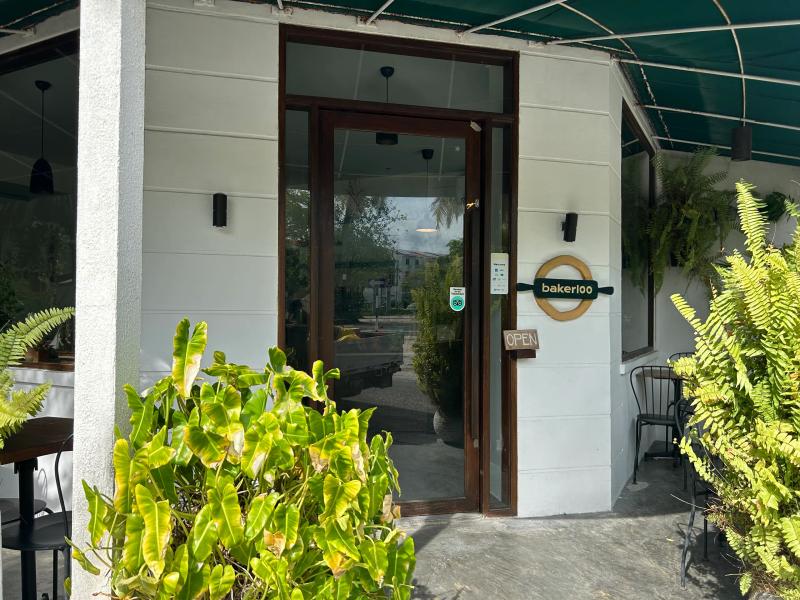 Bakerloo's entrance under a green awning, with a round timber logo by the glass door and ferns either side